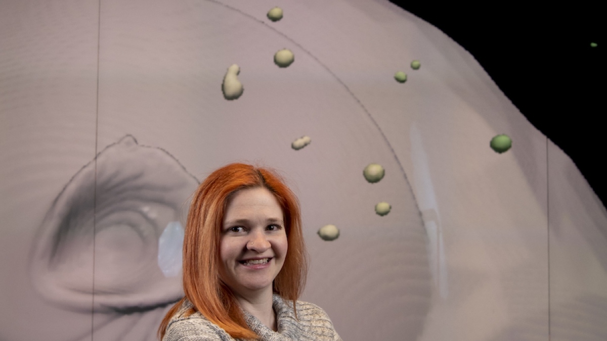 ASU alum Wendy Caldwell stands smiling, with arms folded in front of a digital rendering of an asteroid.