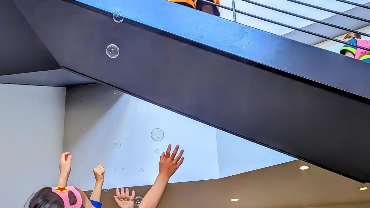 Person stands on staircase blowing bubbles and children below reach up to catch them.