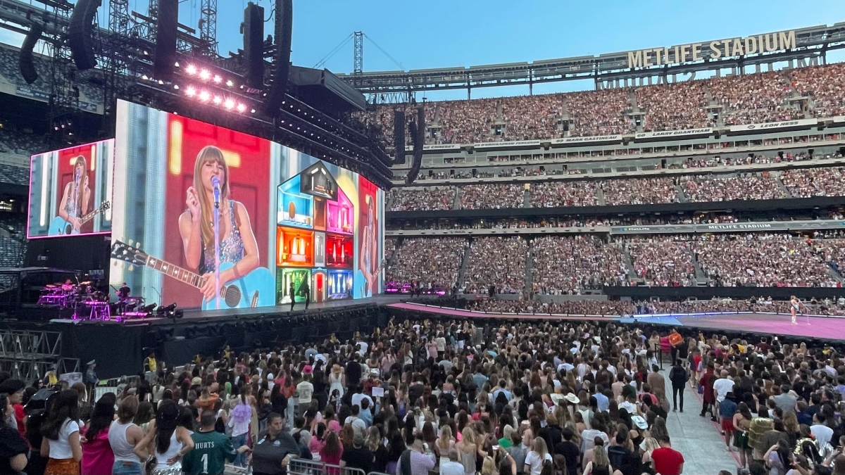 Fans fill a large arena in which a jumbotron displays the image of American singer-songwriter Taylor Swift.