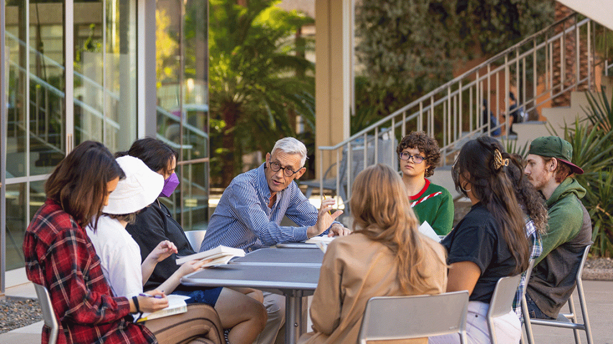 Professor seated at an outdoor table with students listening to him speak.