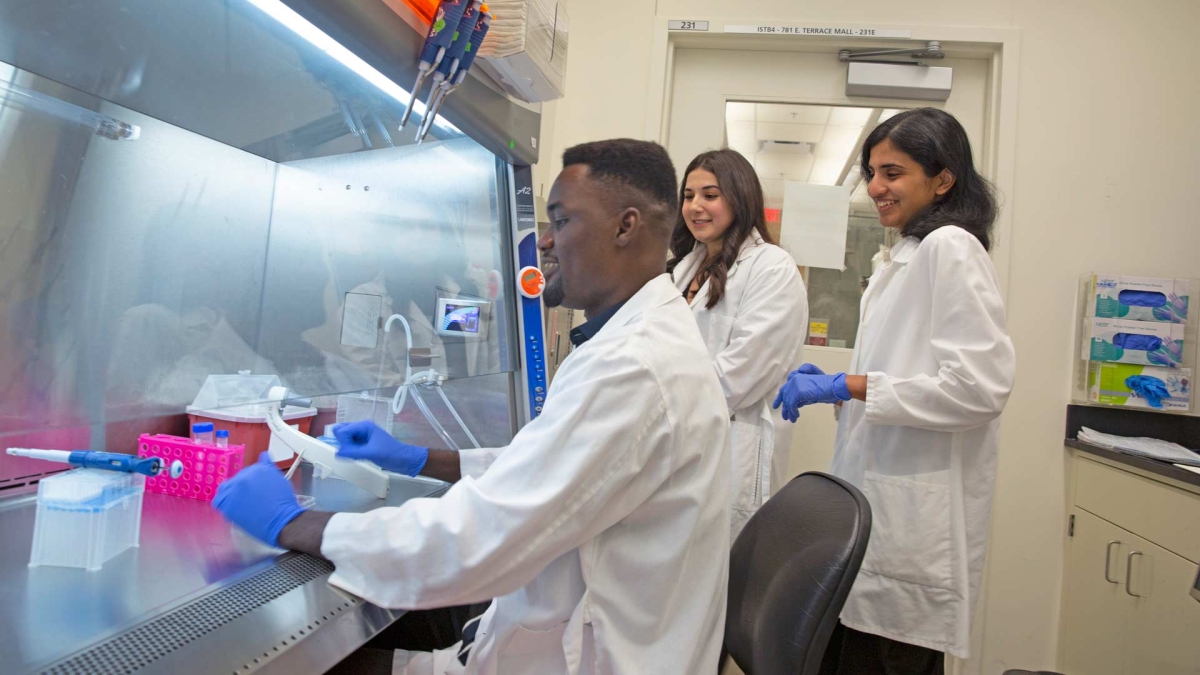 Graduate students Albert Essuman, Gayathri Srinivasan and Carlye Frisch in a lab wearing lab coats.