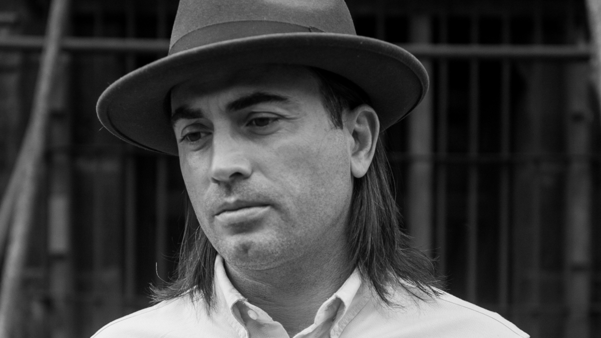 Black and white photo of A.S. Dillingham who has shoulder-length dark hair, a short-brimmed hat, and is wearing a white button-down shirt.