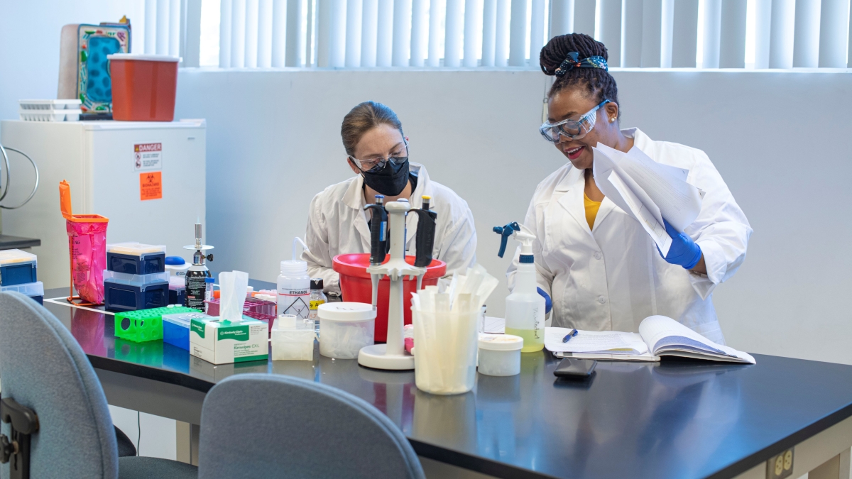 Two people wearing white coats standing at a table in a lab.