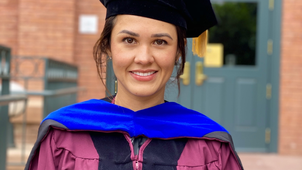 Fall 2020 doctoral graduate Cindy Bonilla-Cirocco stands in front of an old brick building. She is wearing her graduation regalia, including a maroon and black gown, blue hood, and black cap.