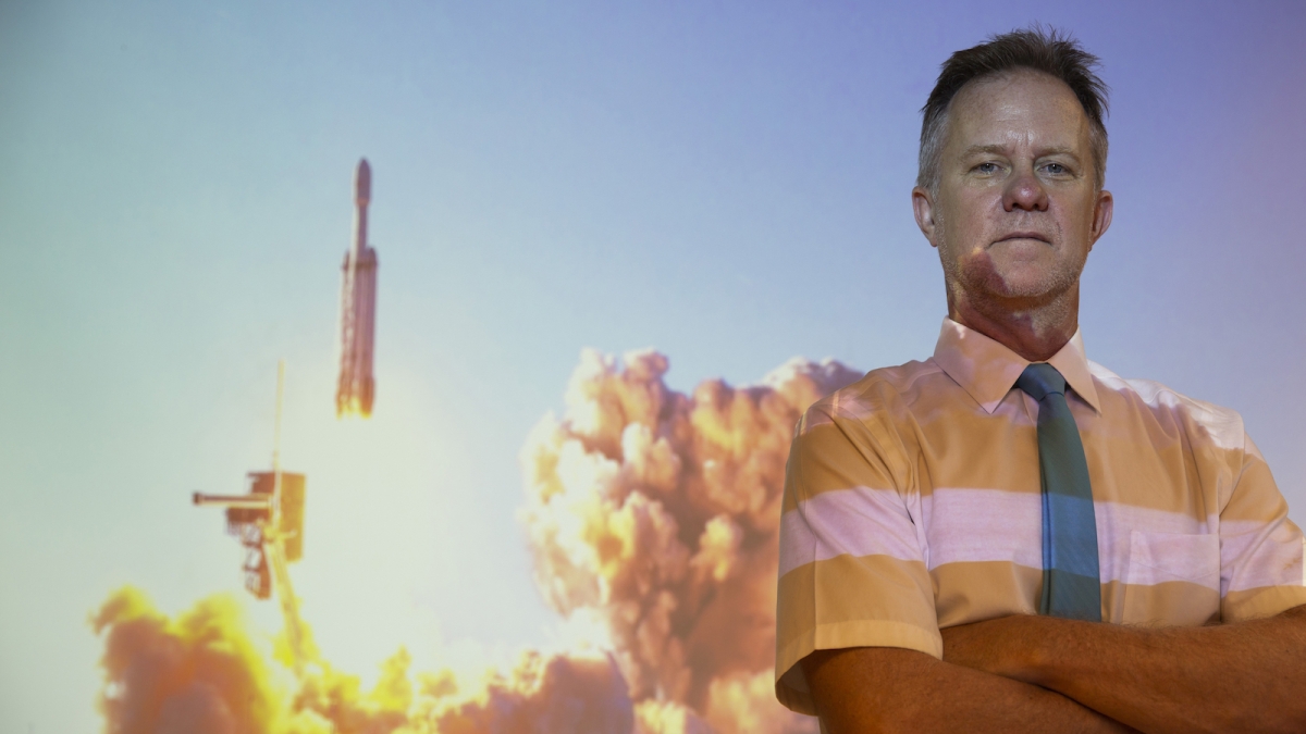 Hugh Barnaby stands in front of a projection of a SpaceX Falcon Heavy rocket launch. 