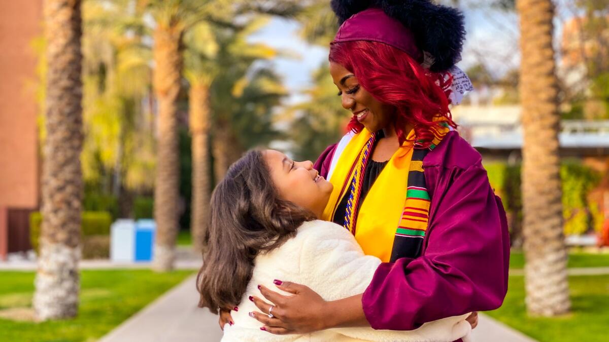 ASU grad Edeline Plaisival in her cap and gown on Palm Walk smiling at her 8-year-old daughter