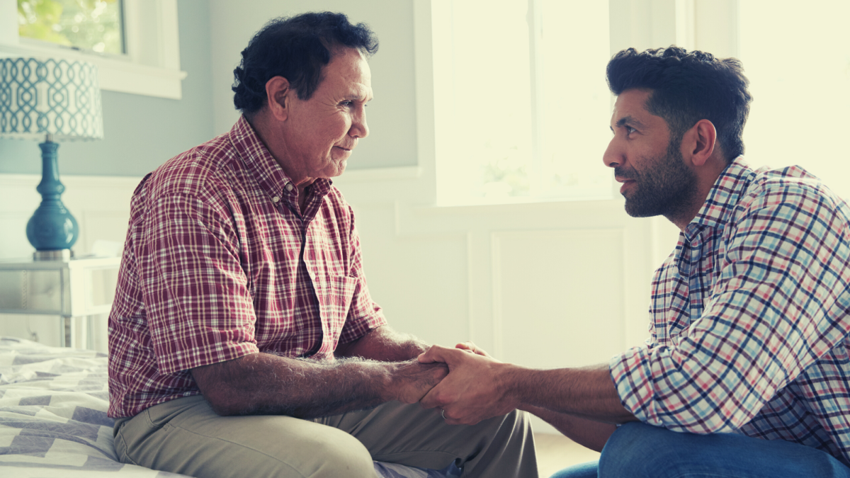 A son who serves as a family caregiver for his elder father, holds his hands as they look at each other. Both men are wearing plaid shirts with bold colors.