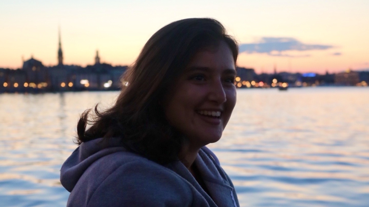 Foreign exchange student Elisa Cardamone is seen in front of a body of water. The sun is low on the horizon and a lit-up city skyline is silhouetted in the distance behind her.