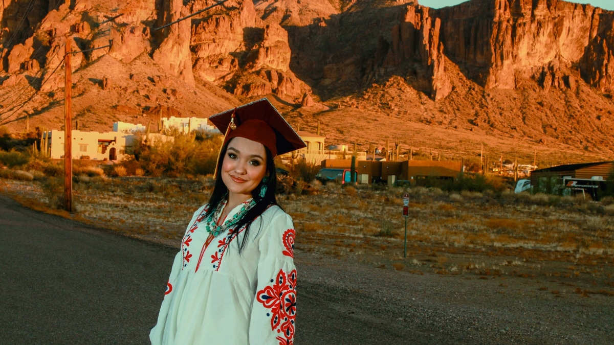 Esperanza Biggs smiles at the camera wearing her ASU graduation cap and a white shirt with red flowers on the sleeves. A mountain range can be seen in the background