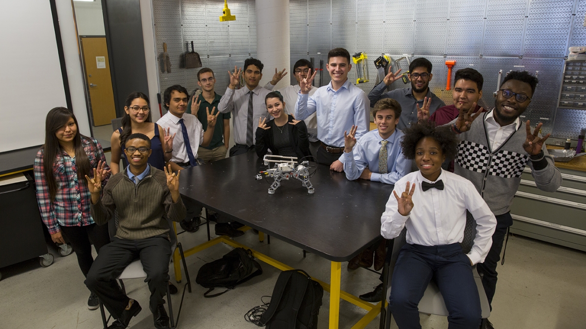 A group of ethnically diverse students gather around a table, displaying the "pitchfork," Arizona State University's signature hand gesture. On the table sits a LEGO robot, and the students are surrounded by tools and equipment.