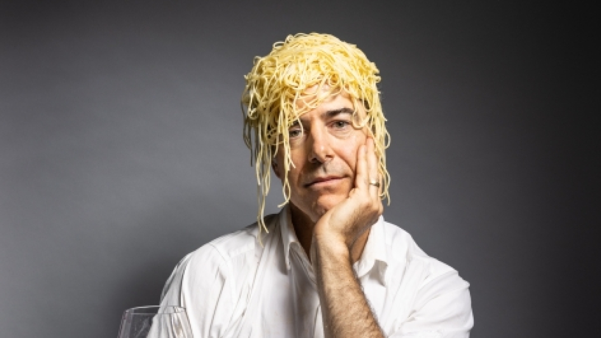 Man with spaghetti noodles on his head seated at a table full of food.