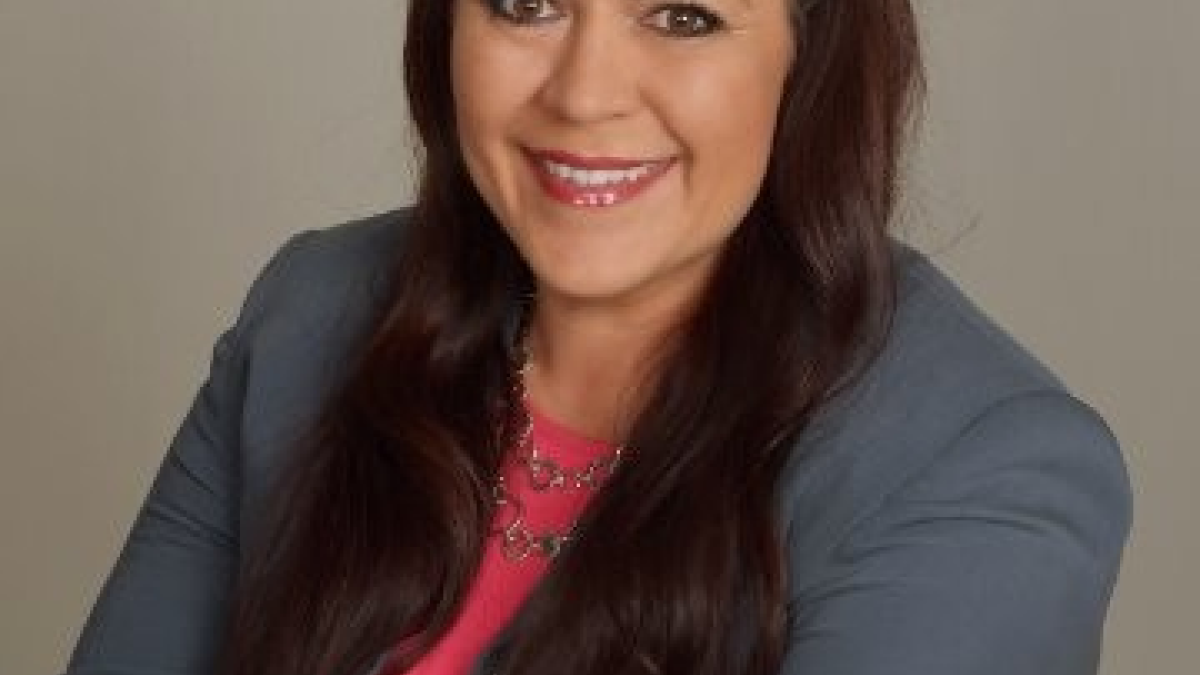 A brunette woman in a grey blazer poses with her arms crossed.