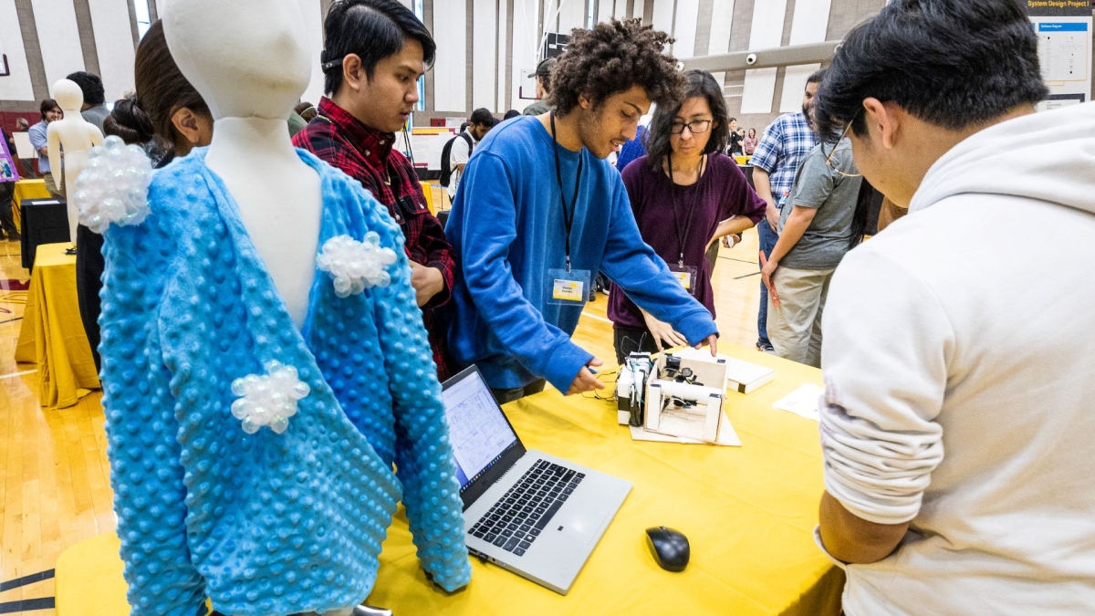 Students gathered around a table with a laptop on it next to a mannequin wearing a blue sweater.