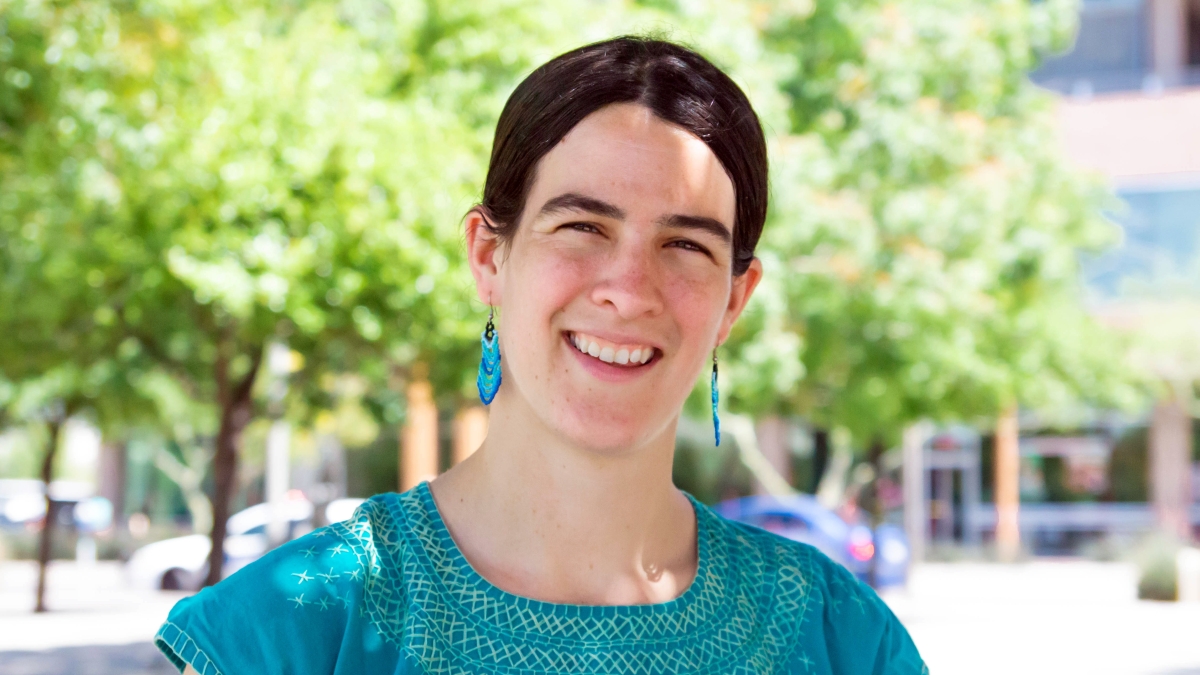 Joanna Williams (light complexion, dark brown hair, wearing teal embroidered blouse and teal dangle earrings) smiles in front of Downtown Phoenix campus
