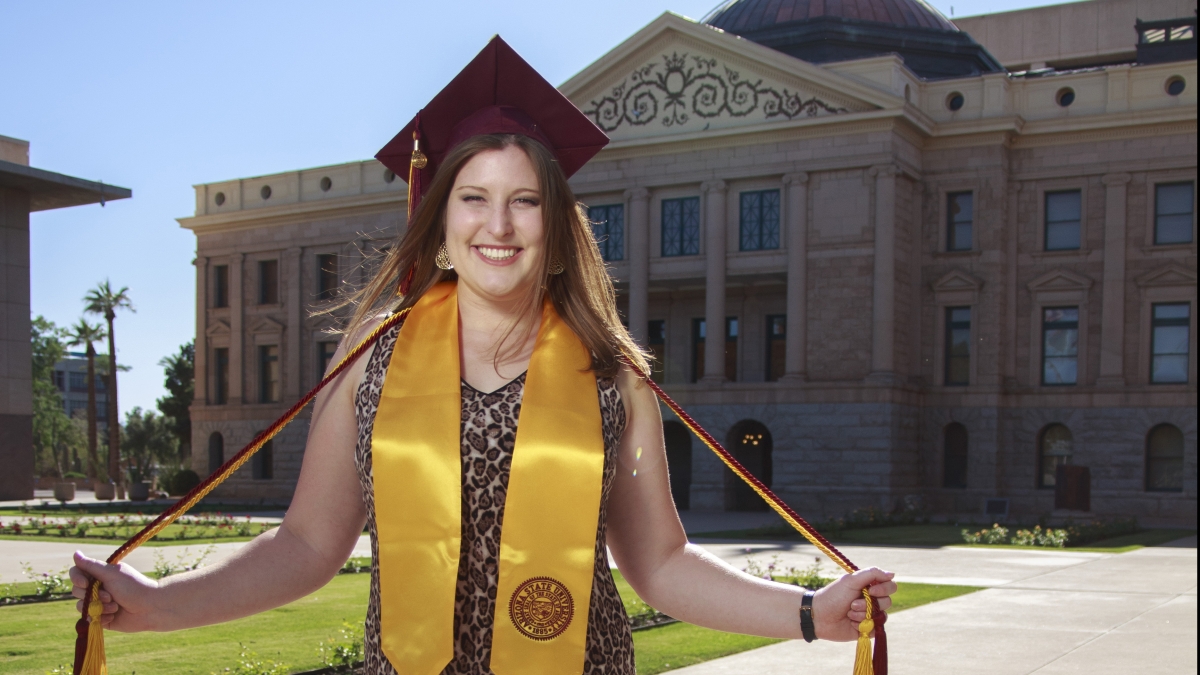 Aly Perkins (light complexion with medium brown hair, wears sleeveless dress, maroon mortar board, maroon and gold cords, and gold stole of gratitude) stands smiling in front of Arizona Capitol building