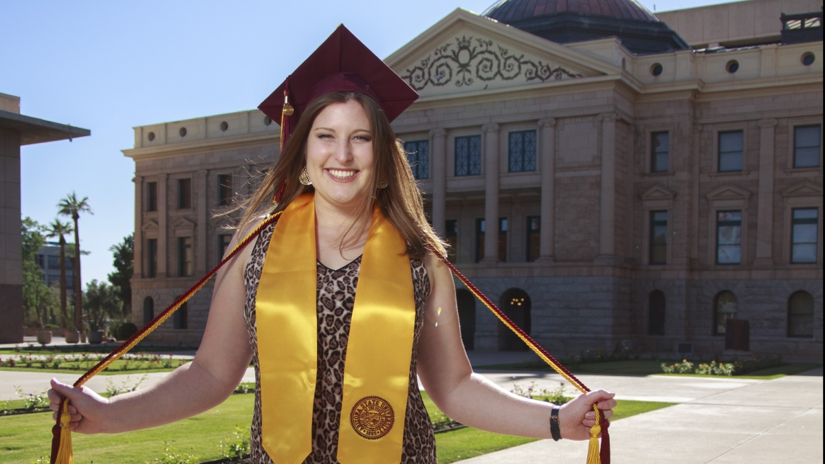 Aly Perkins (light complexion with medium brown hair, wears sleeveless dress, maroon mortar board, maroon and gold cords, and gold stole of gratitude) stands smiling in front of Arizona Capitol building