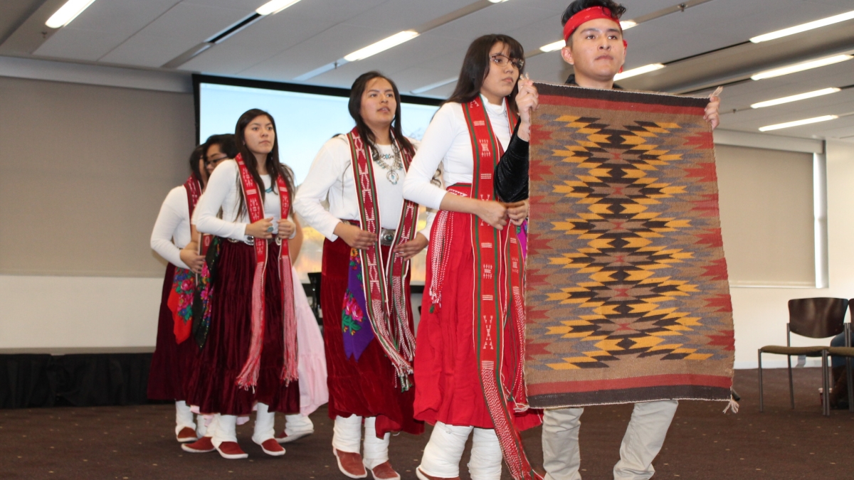 High school students wearing white and red cultural outfits participate in a Language Fair presentation. The students are standing in a line, with the front student displaying a woven rug or tapestry with a red, yellow, and black angular design.