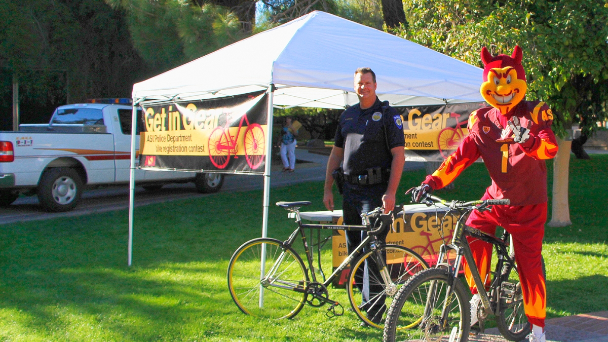 A police officer and Sparky on a bike stand outside near a pop-up tent.