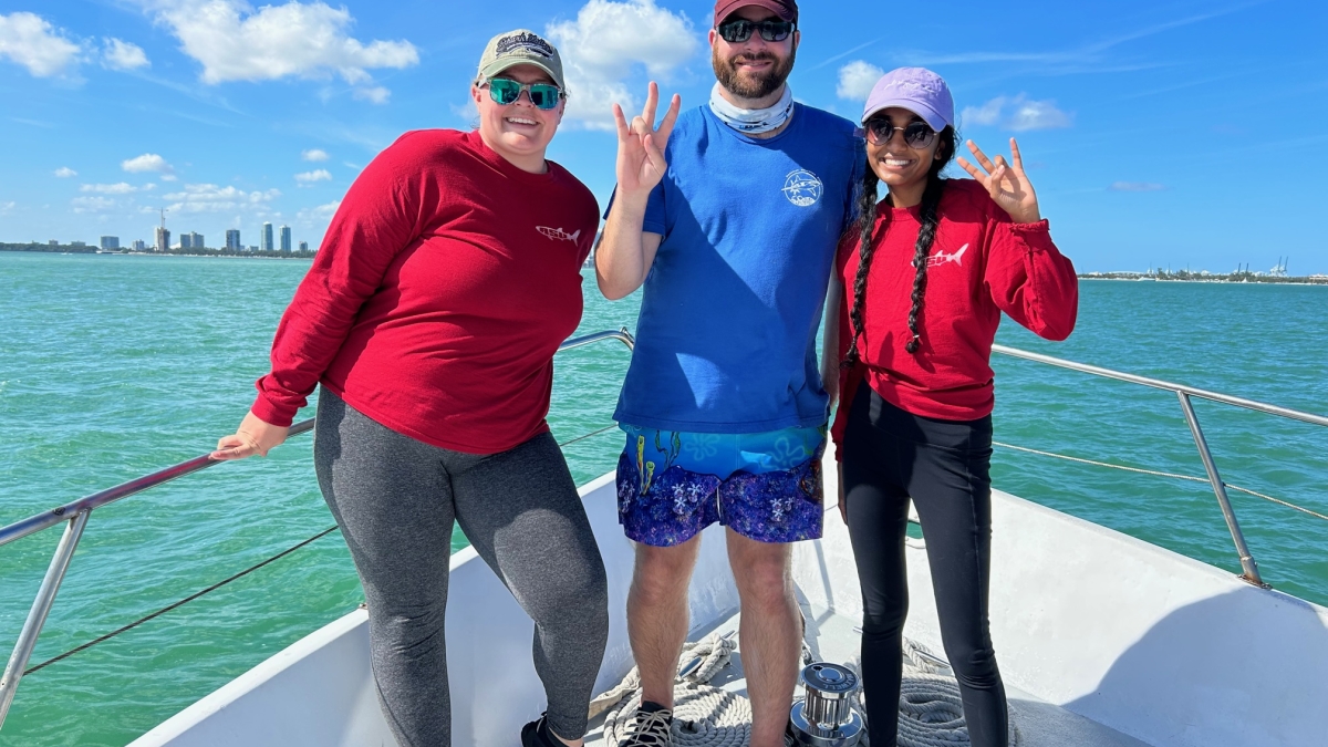 Three people stand on a boat doing the ASU forks up hand sign.