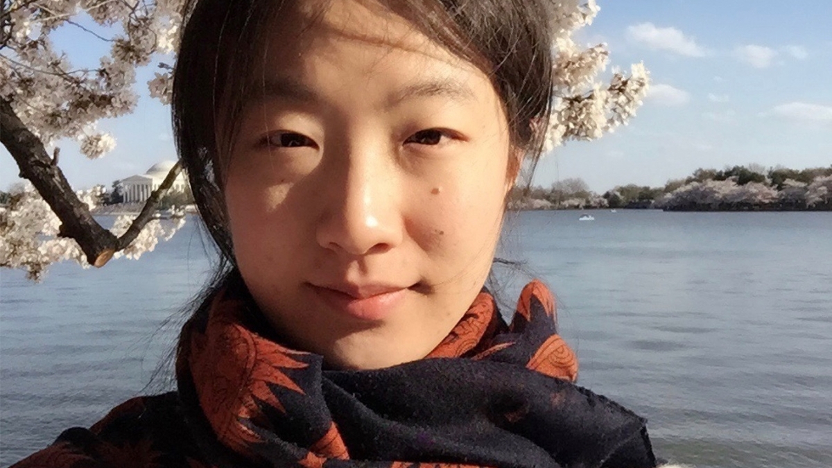 Selfie of Anzi Dong standing in front of a body of water and white cherry blossom tree.