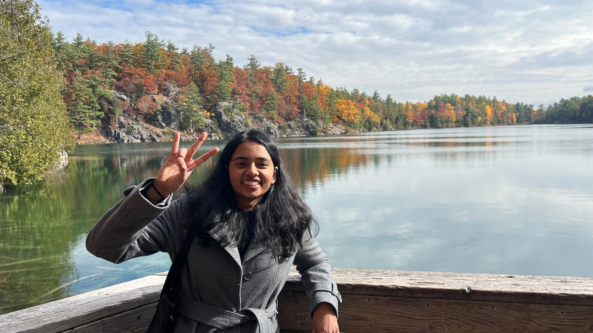 Anusha Natarajan holding up the ASU pitchfork hand sign in Canada, standing on a deck in front of a lake surrounded by trees.