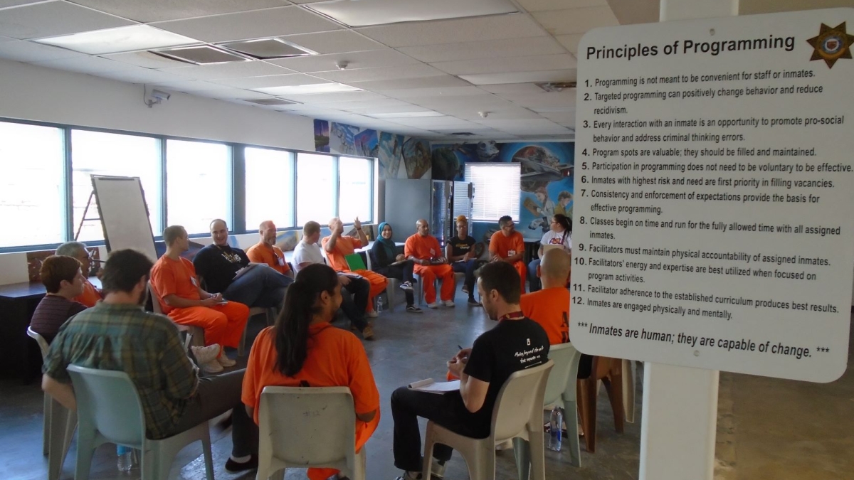 Students and people wearing orange jumpsuits sit on chairs in a circle in a prison classroom.