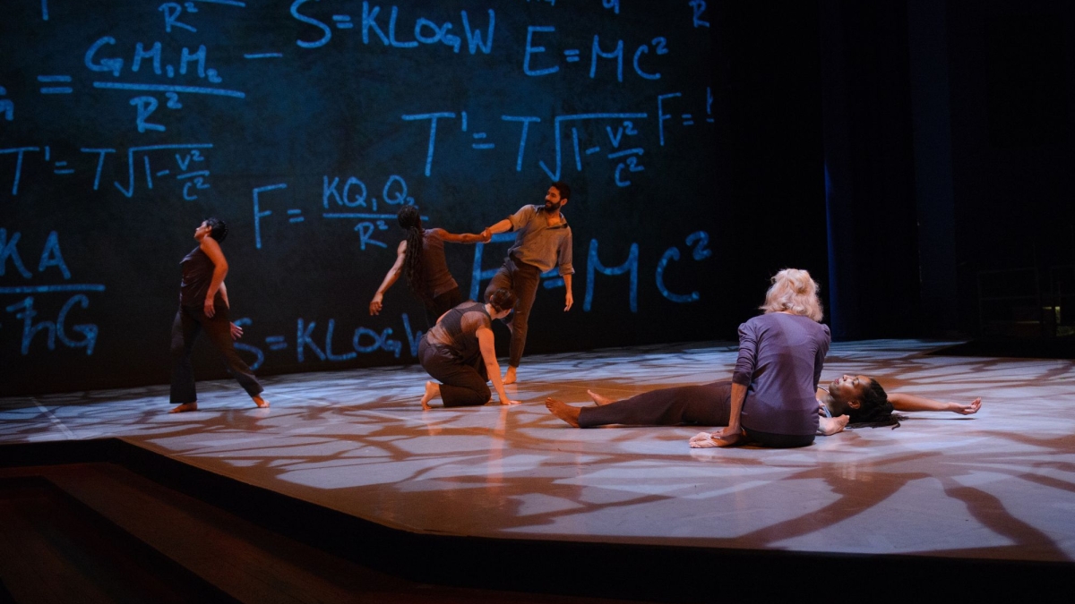 Dancers perform on stage with mathematical equations on a screen behind them.