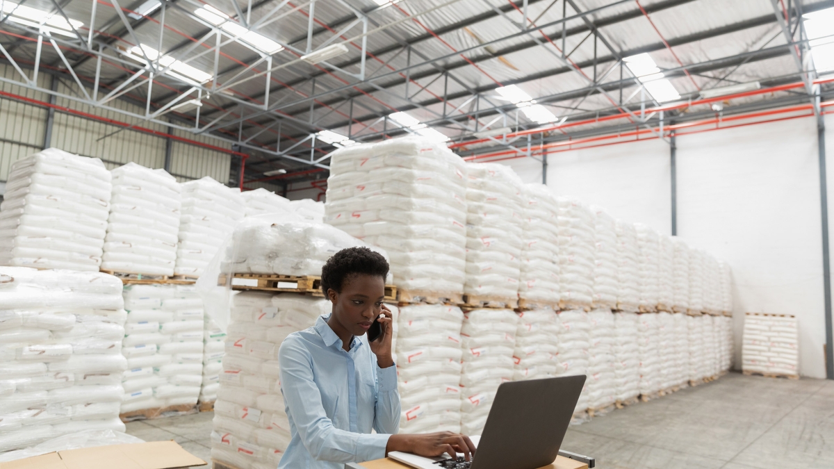 Front view of female manager talking on mobile phone while using laptop at desk in warehouse. This is a freight transportation and distribution warehouse. Industrial and industrial workers concept