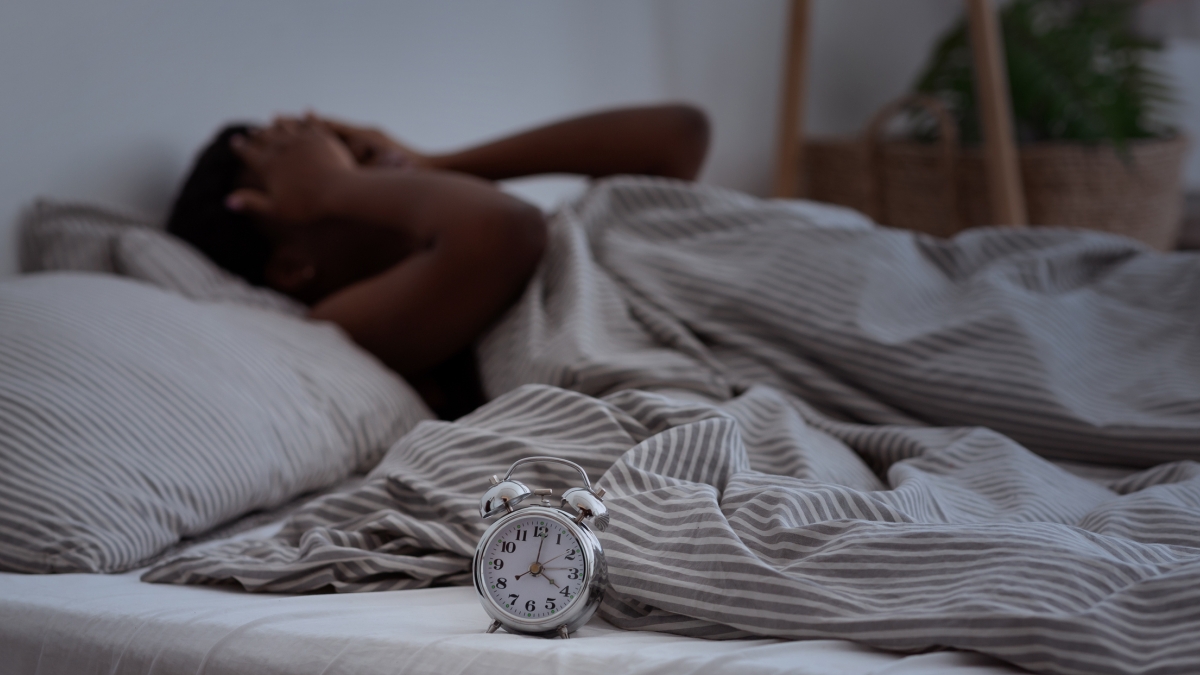 Woman with her hands on her face lying in bed, with an alarm clock next to her.