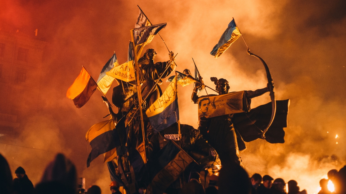 Statues are adorned with Ukraine flags as smoke fills an evening sky around them