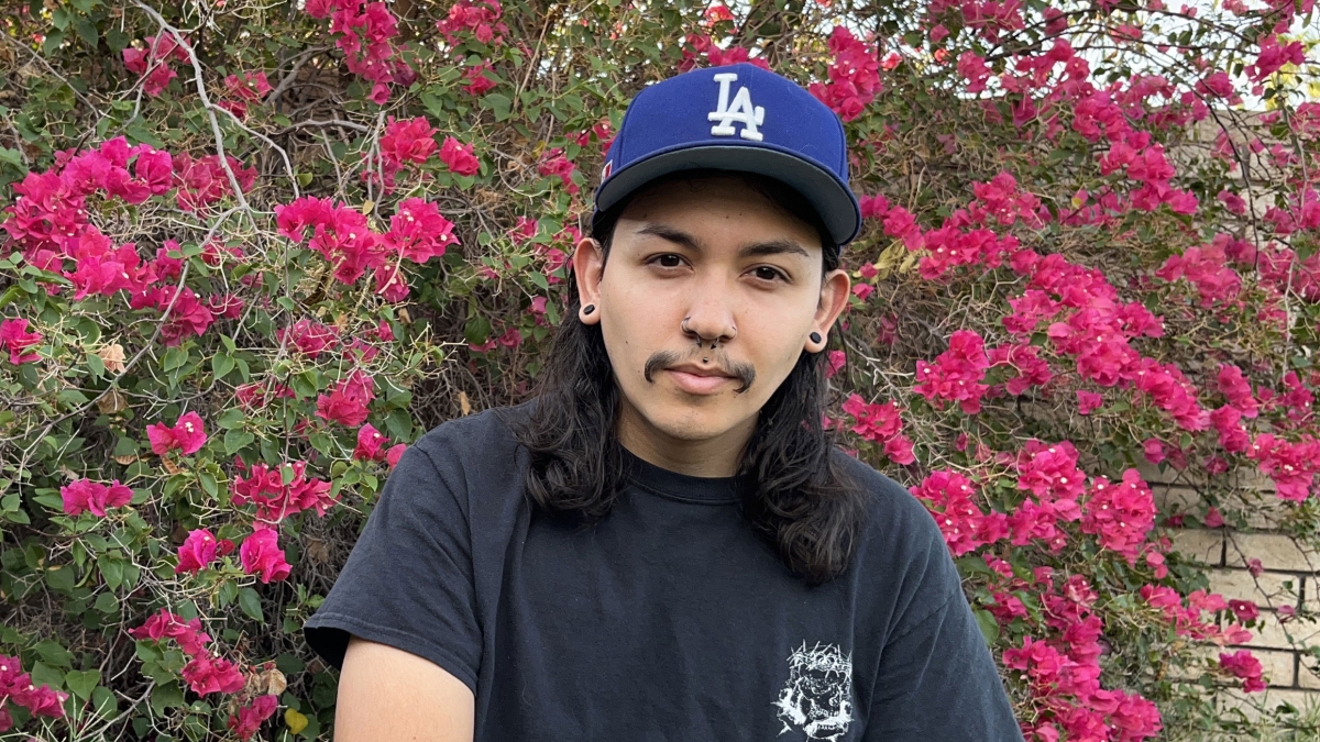 Man with shoulder-length black hair wearing a black T-shirt and jeans and an L.A. Dodgers cap sitting in front of a bougainvillea bush.