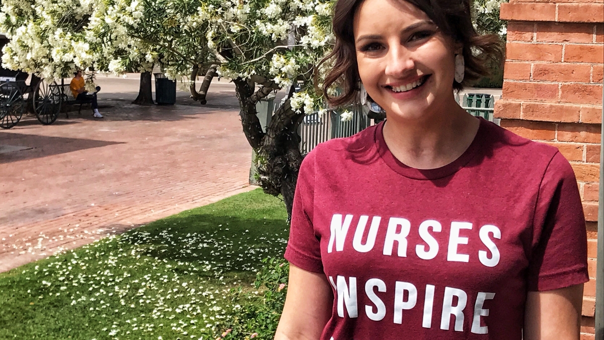 Jaime Buchholz smiles at the camera. She's wearing a nurses inspire nurses shirt and is standing in front of a blooming tree