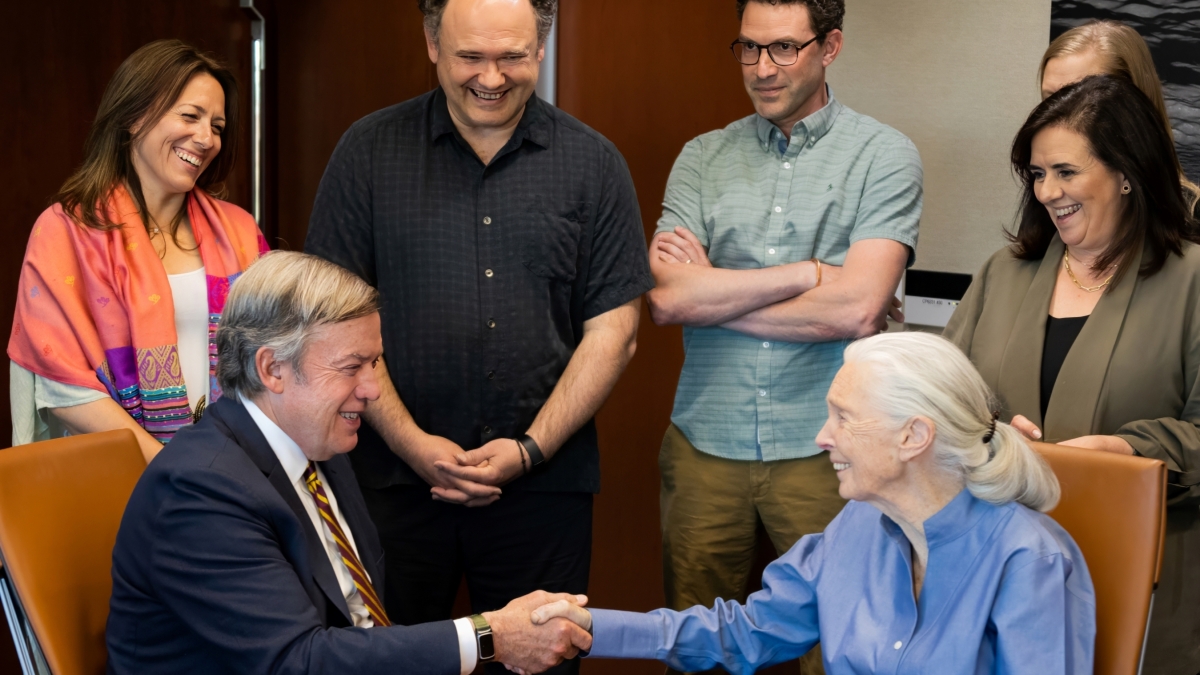 President Michael Crow shakes hands with Jane Goodall while seated at a table with five staff members standing behind them.
