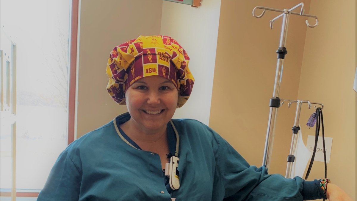 Jennifer Johnson poses in an ASU scrub cap next to an IV machine. She's wearing blue scrubs and smiling at the camera