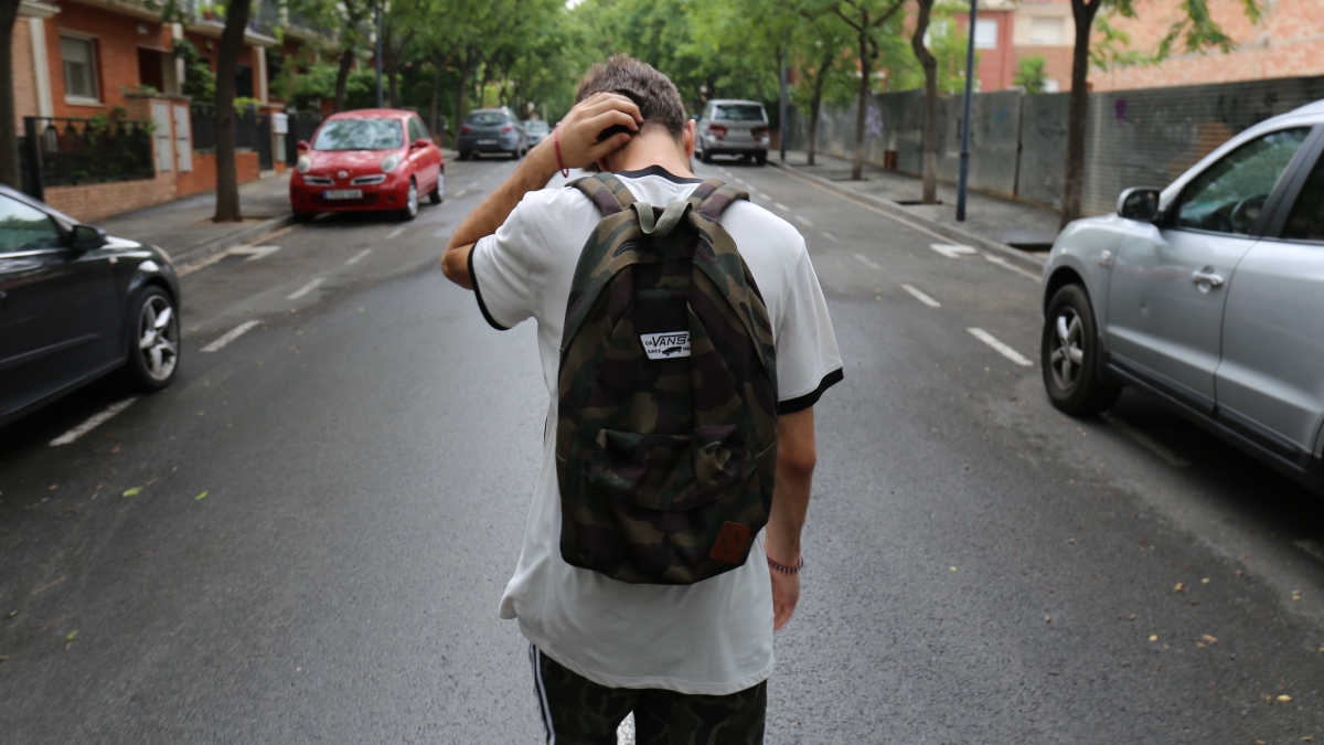 Student seen from behind wearing a backpack and walking down a street while scratching his head.