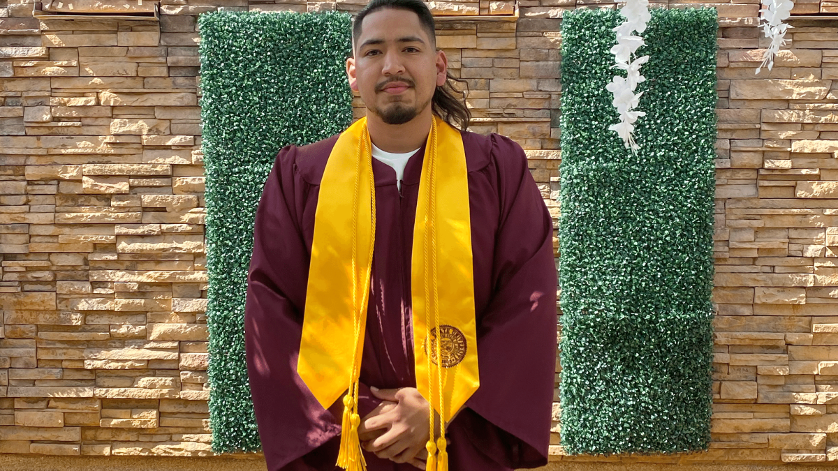 ASU grad Jose Pelagio-Ayala wearing his graduation regalia while standing in front of a brick wall and shrubbery.