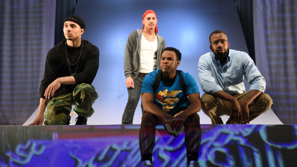 Four young men, some sitting and some standing, in front of a stage-like backdrop.