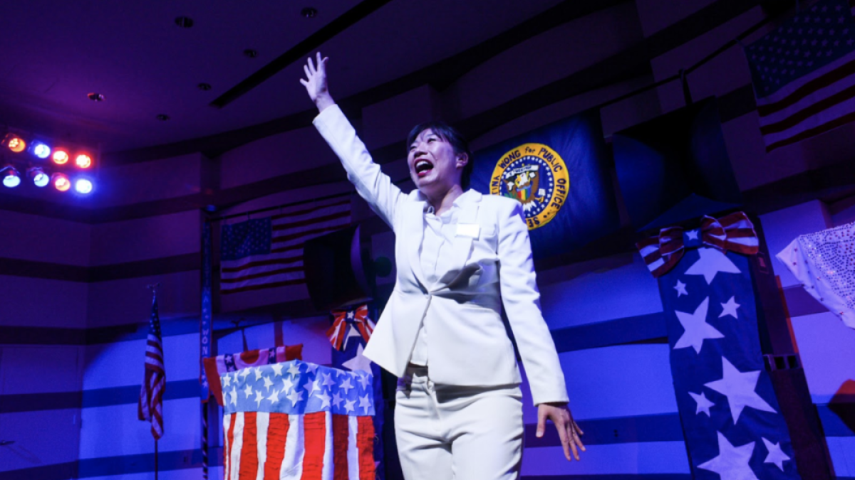 Performance artist, comedian and writer Kristina Wong stanidng on a stage with American flags, wearing a white suit, raising her arm and smiling.