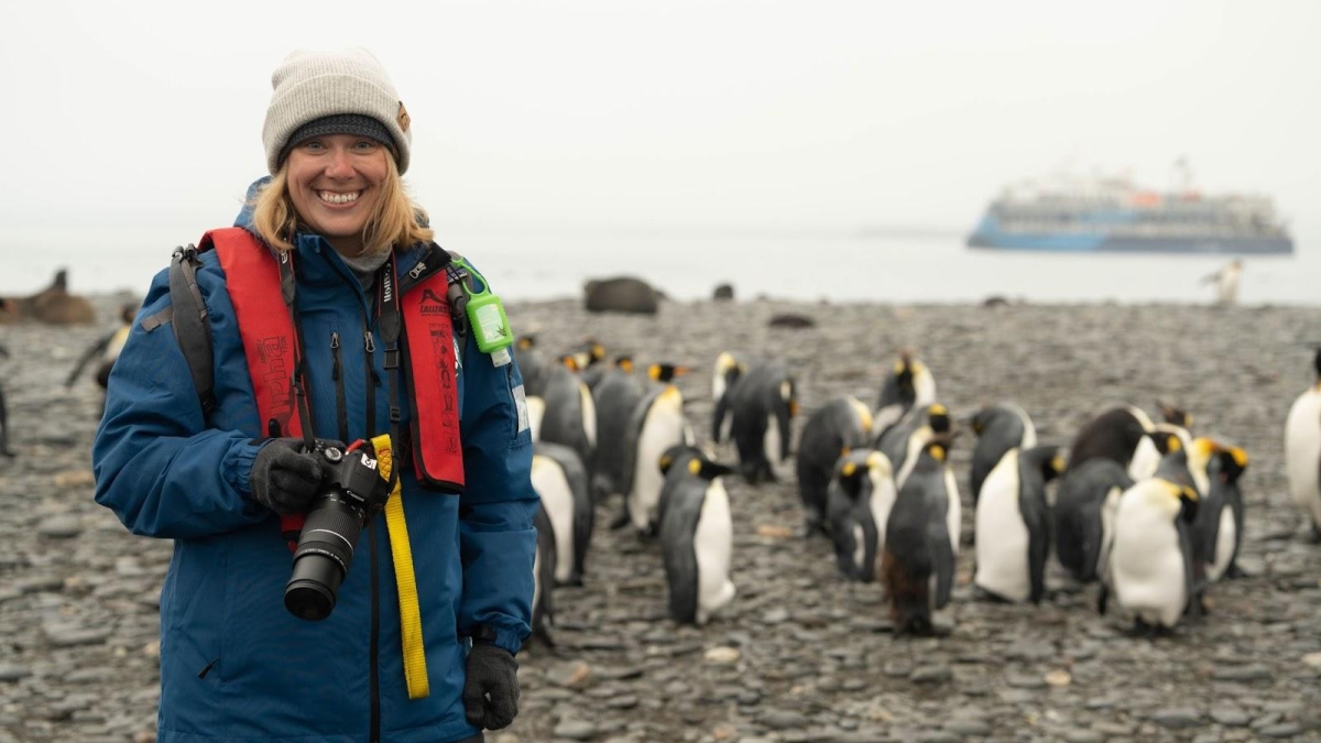 Lauren Richards smiling at the camera in Antarctica wearing a blue parka with penguins and a ship in the background