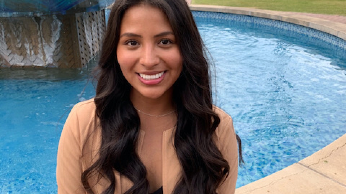 ASU grad Leslie Del Carpio smiles at the camera. She is wearing a black top, a white and black patterned skirt, and a tan blazer. She has long, wavy dark hair. Behind her is a fountain on campus.