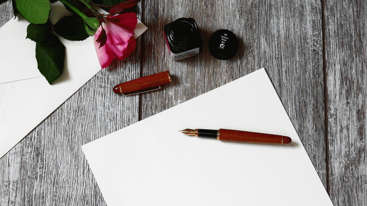 A fountain pen sits on a blank sheet of paper next to an inkwell and a rose