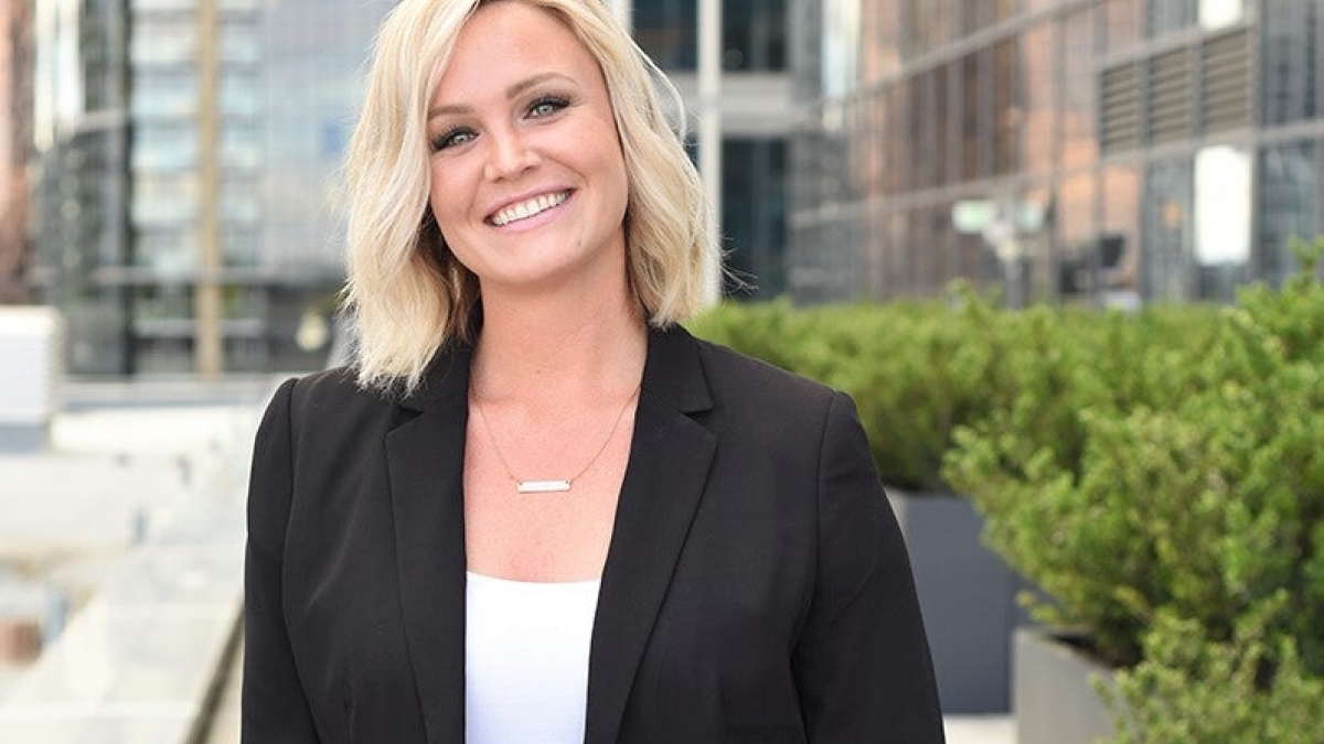 Lindsay Lagreid smiles at the camera. She's wearing a white shirt and black blazer. She's standing in front of a glass office building.