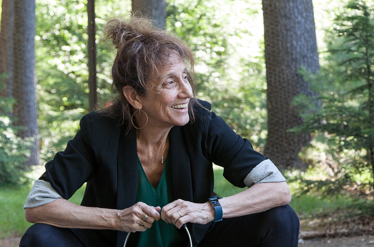 Liz Lerman sitting and looking to the side in a forest-like setting.