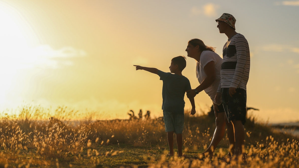 A mother and father stand in a field with a young boy who points toward a setting sun.