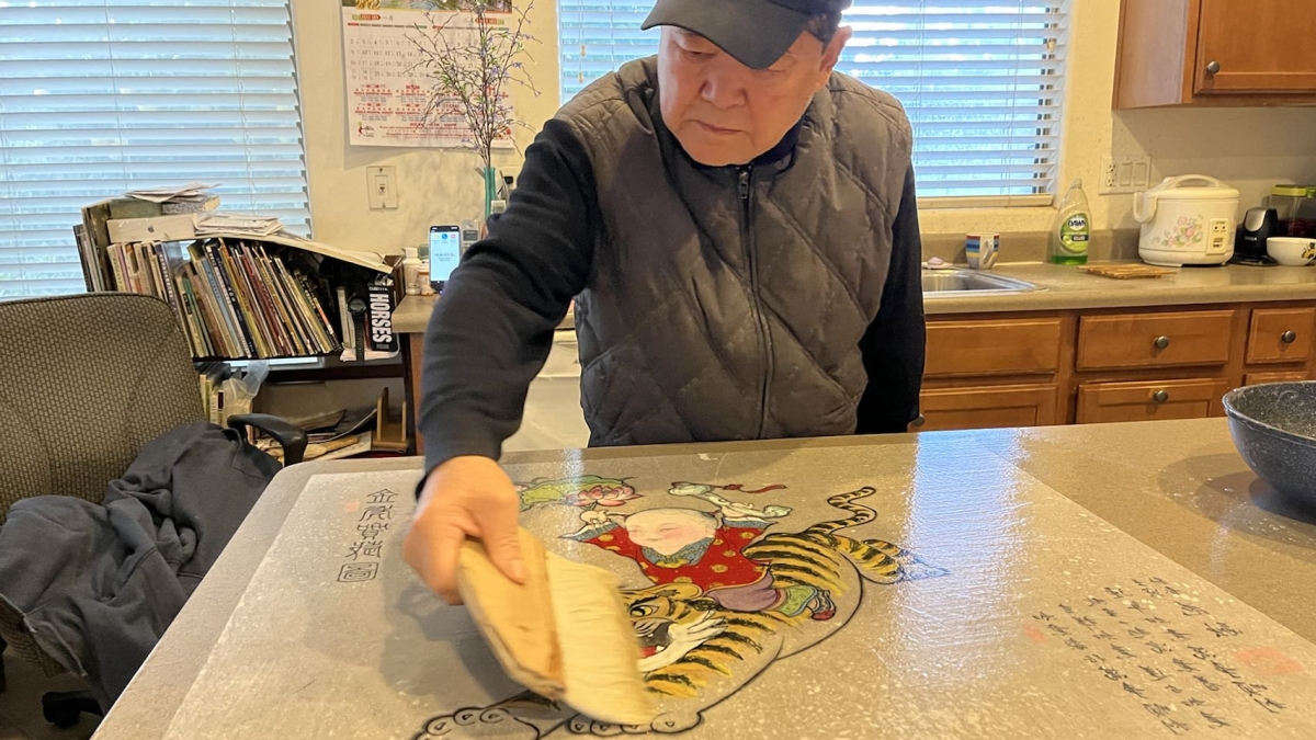 Man painting. He is wearing a gray sleeveless vest over a black long-sleeve shirt with a black baseball cap. He is swiping a wide brush over a painting that is laid across a kitchen counter. 