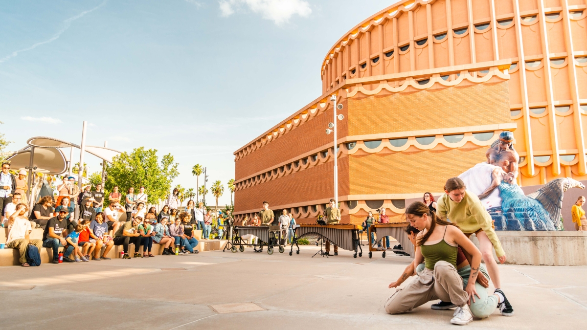 A crowd gathered outside of a circular building watches people perform a dance.