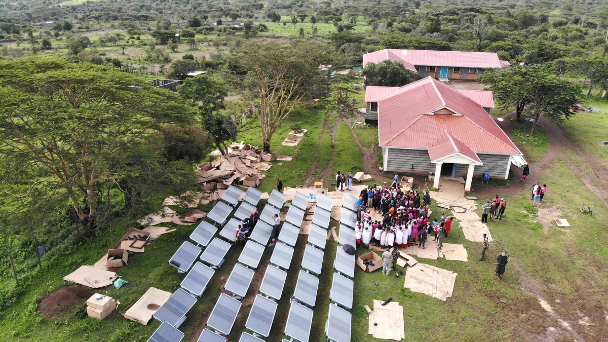 Aerial veiw of a building next to an array of hydropanels in a grassy area. Several people stand near the solar panels.