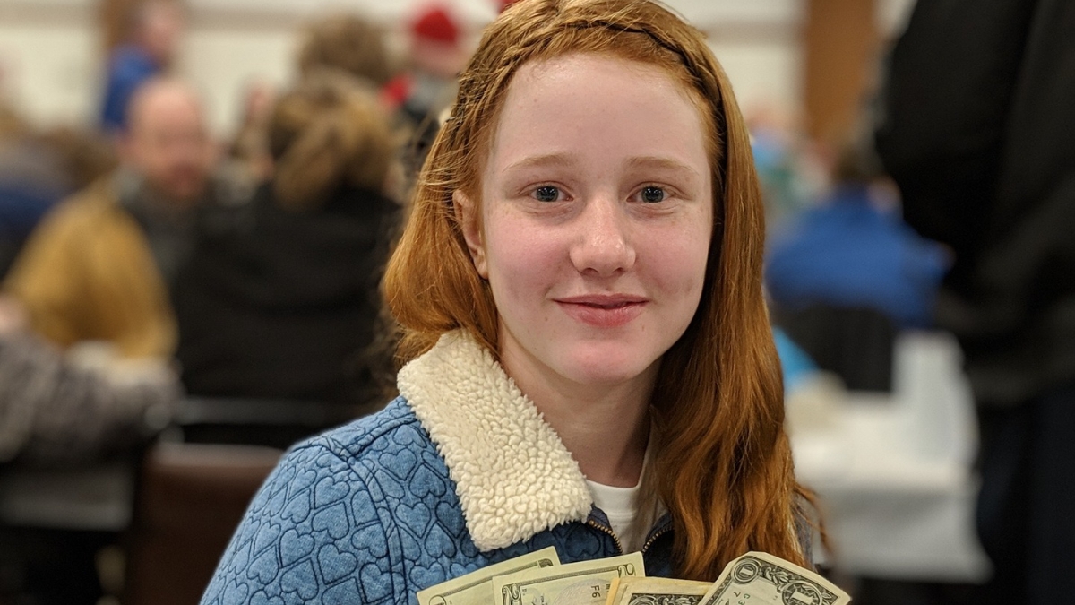 A teenage girl with red hair and wearing a blue coat holds $20 and $1 bills.
