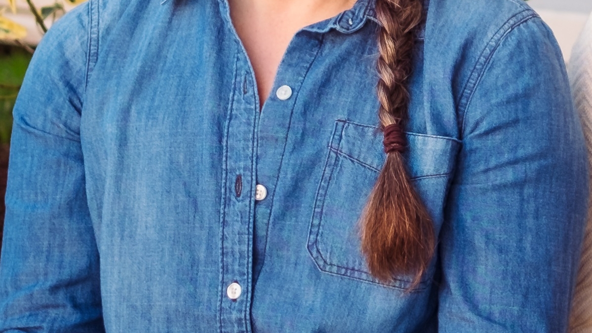 Sabrina Oesterle, a light skinned woman with a long gray and brown braid and glasses, smiles at the camera wearing a blue denim shirt with wrist tattoos showing