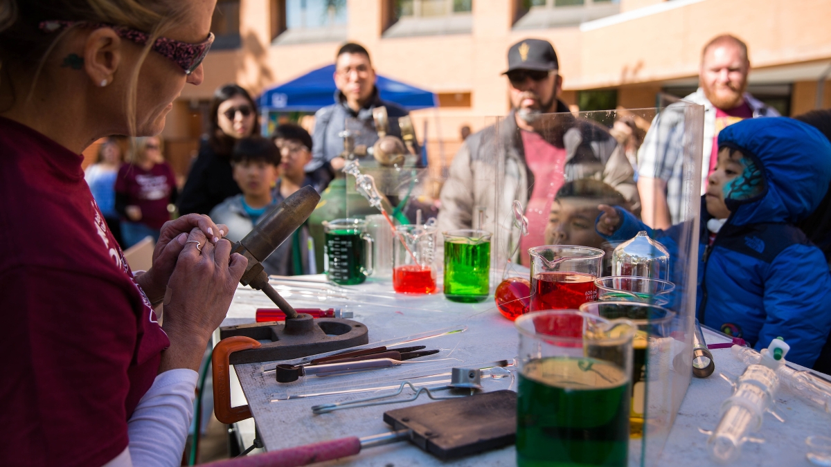 woman holding glass blowing tools with beakers of colored liquid on table while crowd watches